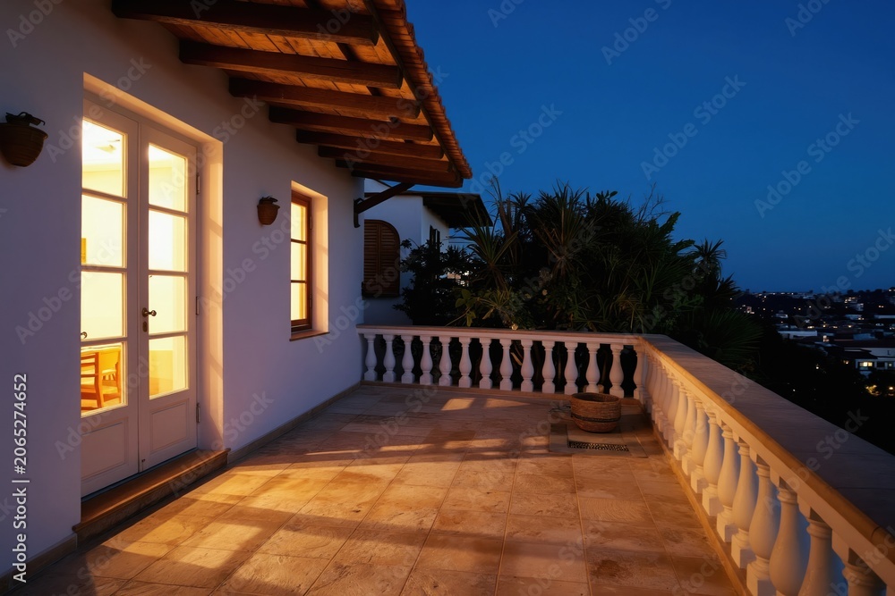 Fototapeta premium Balcony view at twilight with illuminated house windows, balustrade, and distant city lights. Peaceful outdoor space with plants, featuring Mediterranean architecture and warm lighting on tiled floor.