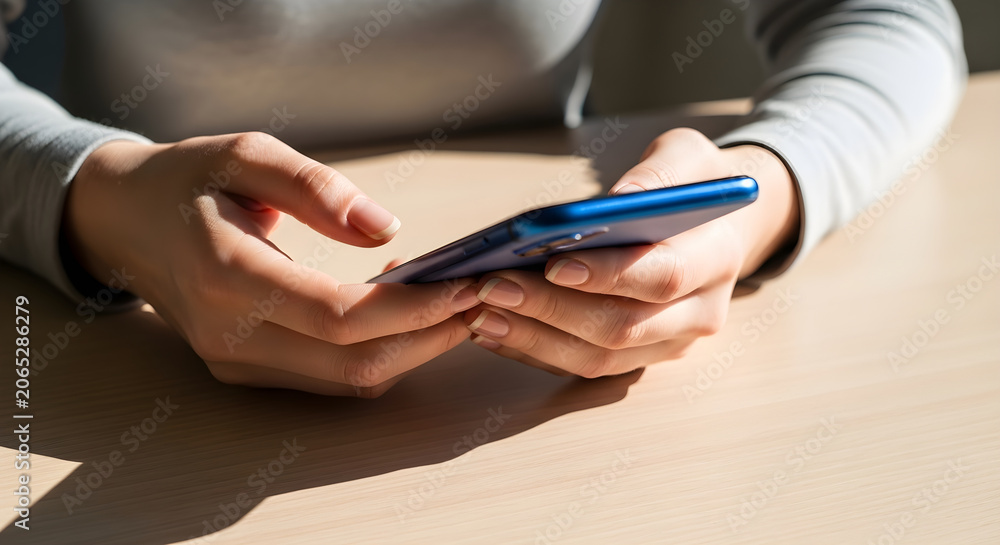 Fototapeta premium Close up of a woman's hands holding and using a blue smartphone over a wooden desk in bright sunlight