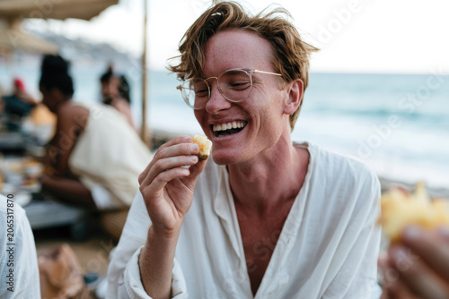A smiling man enjoying a bite of food while surrounded by friends at the beach during sunset, capturing the essence of friendship, fun, and shared moments in life.