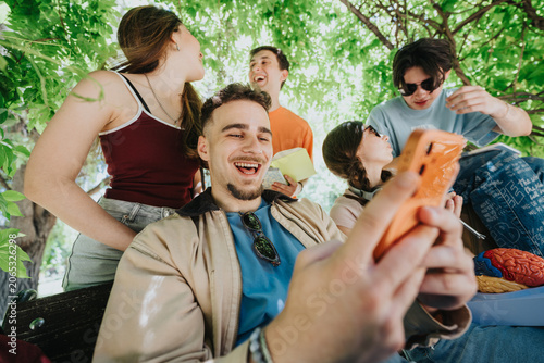 High school students laugh and share a smartphone while sitting under shady trees. A group of teenagers relax together outdoors, studying and socializing in a park-like setting.