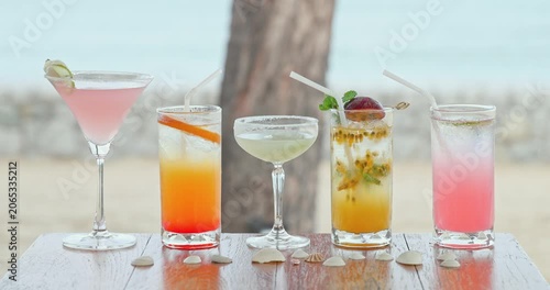 Multi-colored tropical cocktails standing on wooden table with seashells, blurred beach and palm tree background, summer vacation travel promotion, hospitality marketing advertisement
