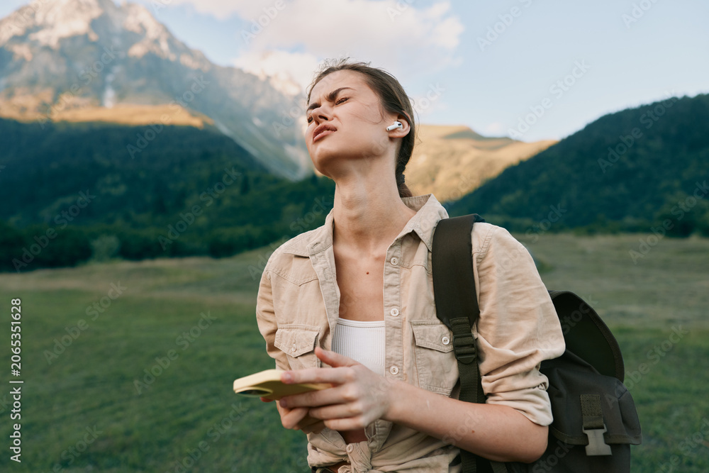 Obraz premium Young woman traveler in a green valley, notebook in hand, backpack over her shoulder, gazing toward distant mountains. Natural light highlights outdoor exploration and curiosity.