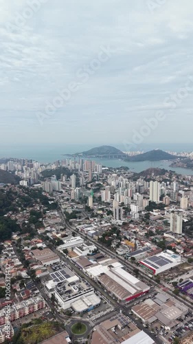 Panoramic aerial view of Vitória, Espírito Santo, Brazil, showcasing the urban skyline, bays, islands, hills, and coastal landscape beneath a dramatic cloudy sky.