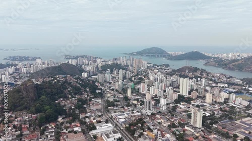 Panoramic aerial view of Vitória, Espírito Santo, Brazil, showcasing the urban skyline, bays, islands, hills, and coastal landscape beneath a dramatic cloudy sky.