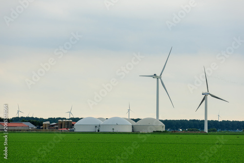 A modern biogas plant featuring white dome-shaped storage tanks paired with industrial wind turbines in a lush green agricultural field