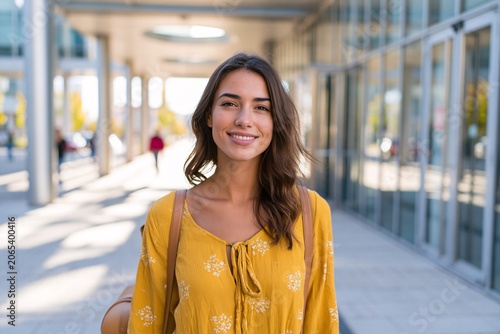 Smiling woman in a yellow patterned dress stands outdoors near modern glass buildings