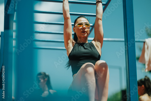 Canvas Print A fit woman performs hanging leg raises on outdoor monkey bars while wearing sunglasses