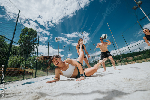 A group of young friends play beach volleyball as a woman dives in the sand to reach the ball. The outdoor game shows energetic action, laughter and summer fun on a fenced sand court.