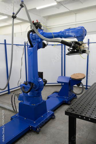 Blue articulated robot arm stands ready in clean industrial cell. Flexible extraction hoses and abrasive disc fixture highlight automated material processing setup.