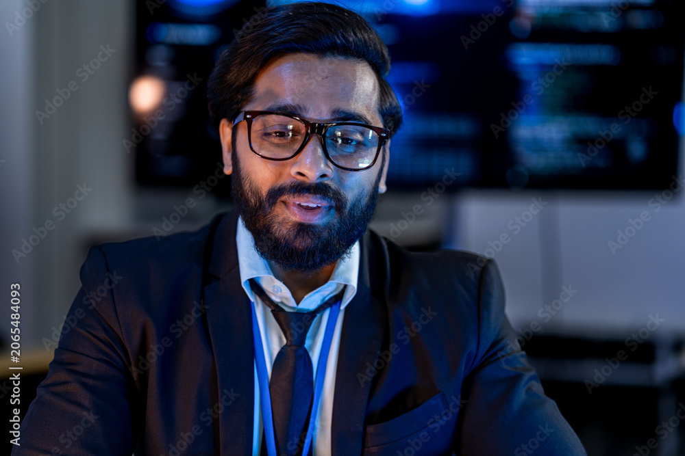 Obraz premium South Asian male network security engineer in a dark blazer and blue badge presents attack vector analysis in a dark room with blue-lit threat dashboards.