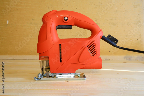 Bold red electric jigsaw stands poised on light wood surface. Workshop energy meets clean composition for carpentry and DIY projects