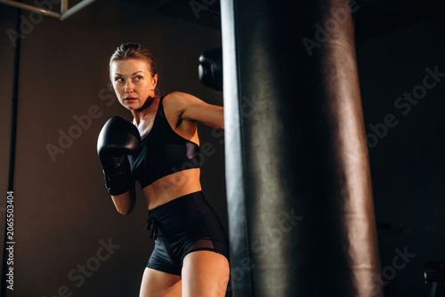 Obraz na plátně Female boxer in black sportswear practicing punches against a heavy bag in a dim