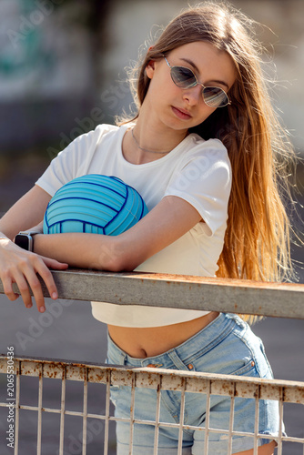 Outdoor portrait of a blonde pre-teen girl wearing bright blue sunglasses and holding a matching blue ball