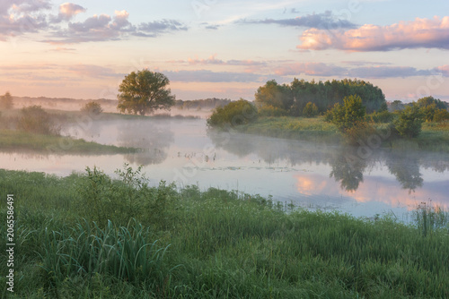 A peaceful river landscape at dawn with thick morning mist over the water, lush green banks, and soft sunset clouds