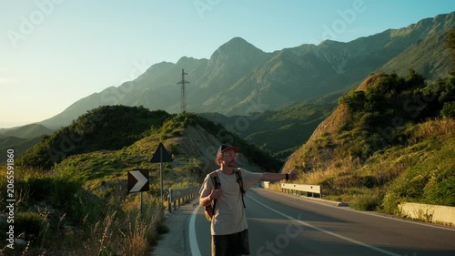 Handheld wide shot of male backpacker on mountain roadside signaling for ride, lifting cap to flag down a car. Warm sunset light, open road and waiting pose highlight hitchhiking and urgency.