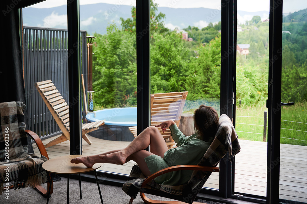 Fototapeta premium Woman relaxing in armchair with legs on table, enjoying scenic mountain view through large window, modern cozy cabin interior with terrace, outdoor hot tub and wooden lounge chairs