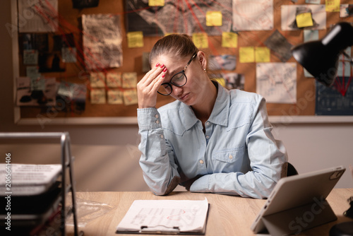 Female detective or police investigator holding head with hands, feeling stressed while working late at night in office