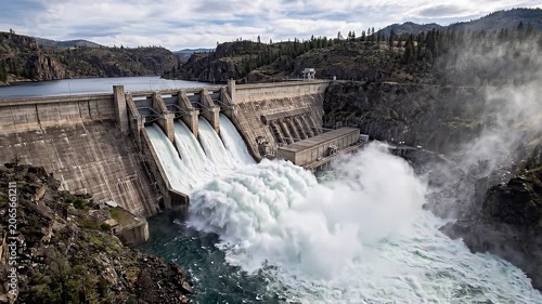 Aerial tracking shot of dam spillway releasing massive water flow into rocky canyon reservoir under overcast sky with mist and spray