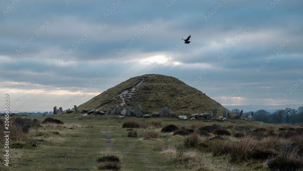 Fototapeta premium Ancient archaeological site with a grassy hill, standing stones, and a bird flying in cloudy sky