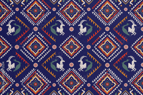 Seamless textile pattern featuring stylized ibex figures, intricate diamond shapes, and floral motifs in blue, orange, and white colors