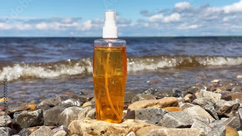 Transparent bottle of tanning lotion stands near water with waves in background