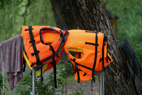  Bright orange life jackets drying on a rack outdoors near a tree.  Detailed close up of orange personal flotation devices with black straps on a background of nature.