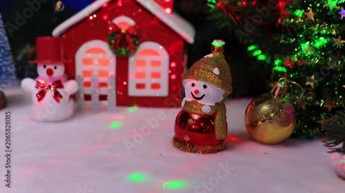 Near the little red house there is a snowman with a golden ball and a Christmas tree and a snowman in a red hat
