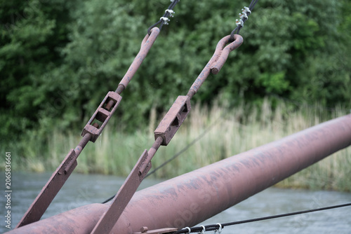 Detailed close up of industrial rigging equipment and rusty steel pipelines. Heavy steel cables and turnbuckles holding a large metal pipe outdoors.