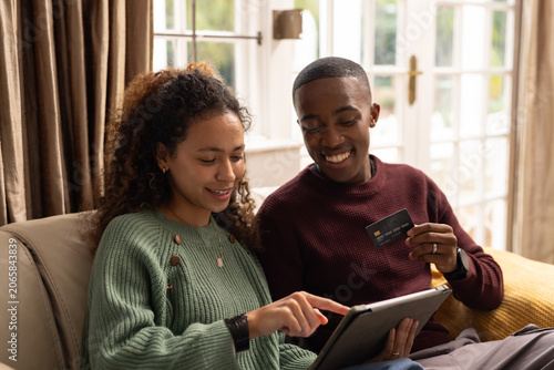 Couple sitting on beige sofa at home, wearing knit sweaters and using tablet with credit card