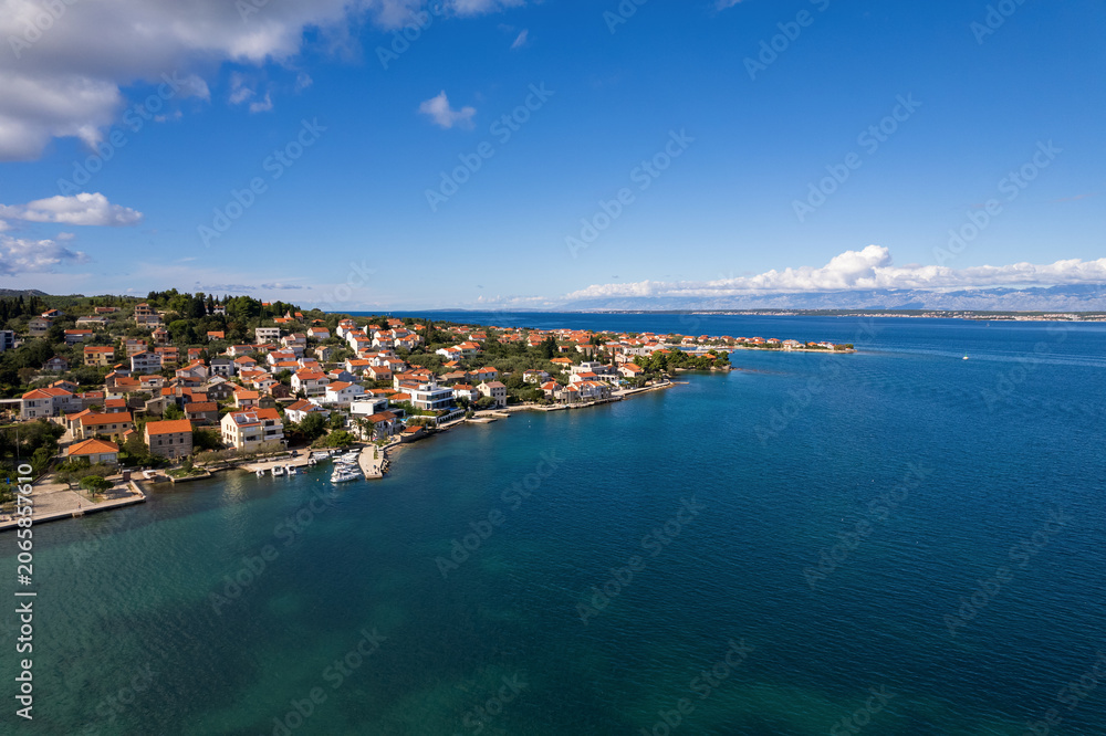 Obraz premium Drone shot capturing the beautiful Adriatic sea coast looking towards Poljana village from Preko side on Ugljan island, Croatia. Red-roofed houses line the turquoise water bay under a blue sky.