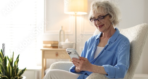 Canvastavla Senior woman with smartphone in armchair at home. Space for text