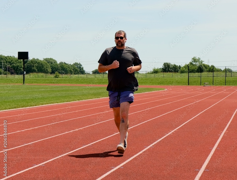 Fototapeta premium Adult man preparing for outdoor workout adjusting wireless earbuds stretching legs on railing and running on a red running track at a local stadium