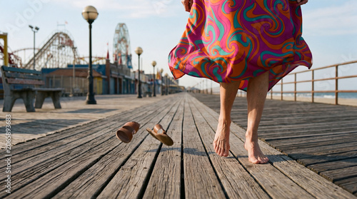 Senior woman feeling free and carefree, walking barefoot on a wooden boardwalk, her colorful dress and flying sandals adding to the mood