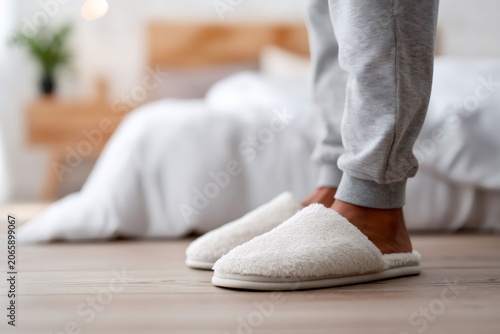 Man wearing soft white slippers standing on wooden floor in bedroom. Low section of male legs in sweatpants next to bed. Morning routine and comfort concept