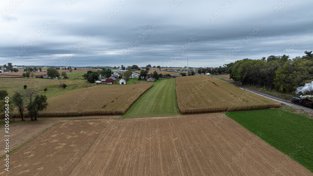 Naklejka premium Cornfields line the land with a few houses visible in the distance. A railway track runs along the edge of the fields as clouds fill the sky. The scene shows rural life on a fall day.