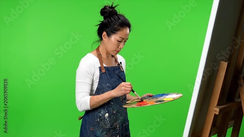 Young woman in a denim apron holding a digital tablet while standing in front of a green screen studio backdrop.
