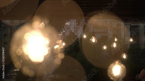 shot cafe set roof building, raindrops falling garland yellow light bulbs, moved wind, car headlights shining background below twilight summer day. blurred raindrops, bokeh warm incandescent lights