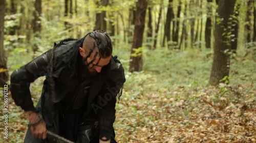 Norse adventurer, Chopping logs, Dark outfit. Dressed in dark clothing, bearded Viking chops timber in forest filled with autumn colors.