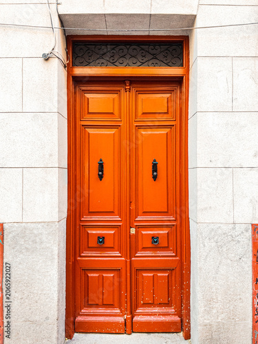 European double wooden entrance doors in a rich orange-red color, set within a stone building facade with black iron hardware.