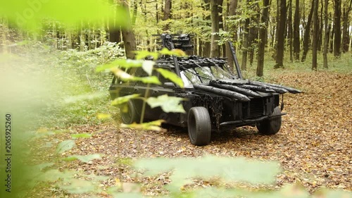 Post-apocalyptic theme, rugged terrain, dense foliage. In wooded clearing, heavily armored vehicle with spikes is partially obscured by leaves, capturing dystopian or post-apocalyptic feel.