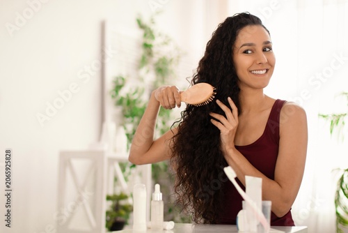 Schilderij op canvas Happy Young Woman Smiling While Brushing Curly Hair With A Wooden Brush In Bright, Cozy Bathroom
