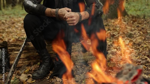 Fallen leaves, Campfire warmth, Campfire comfort. Surrounded by fallen leaves, rugged person in protective gear sits by blazing campfire in forest.