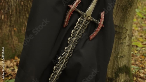 Worn sword, Mystical forest, Cloaked warrior. Person shrouded in dark cloak traverses forest, with worn sword slung over their shoulder.