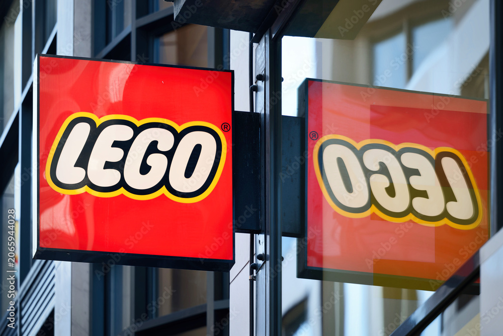 Naklejka premium Cologne, Germany June 18 2026: Red Lego brand sign on the facade of a retail store in Cologne, reflecting backwards into the large glass window pane of the storefront on a bright day