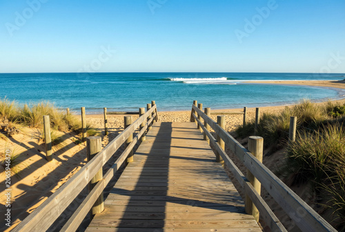 A peaceful landscape featuring a rustic wooden boardwalk path through dunes toward a vibrant turquoise sea with soft waves and gold sand under bright sunlight. Generative AI.