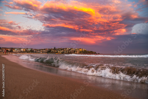 Gentle waves wash onto Bondi Beach beneath a dramatic sunset sky with vibrant cloud formations and the Sydney coastline in the background, Australia.