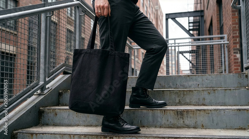 Man in dark pants and boots carries a black tote bag up outdoor metal stairs