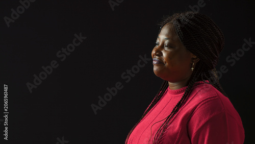Obraz na plátně Positioned against isolated Black backdrop young woman wears downcast facial expression, with side-view portrait of African American fashion blogger in a mournful stance against blank background
