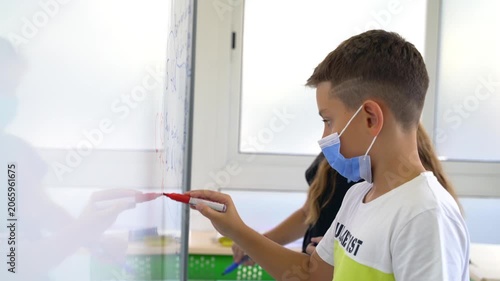 Boy wearing face mask draws on glass wall with marker in classroom