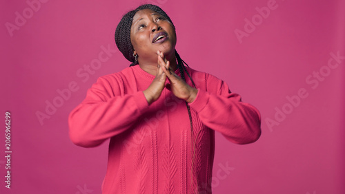 Fotografie Young lady with African American ethnicity standing and praying with faithfulness against pink background, with Black woman in elegant style looking above with her hands up showing devotion